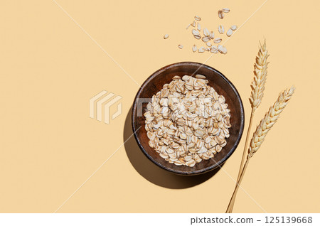 Oat flakes in a wooden bowl with wheat on a yellow background with shadow. 125139668