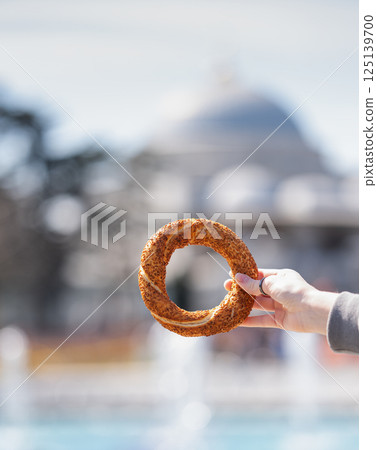 A hand holds a sesame simit outdoors in front of a mosque on a sunny day. A hand holds a sesame simit outdoors in front of a mosque on a sunny day. 125139700