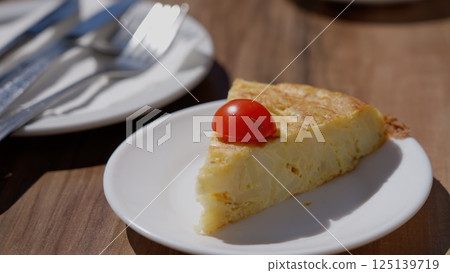 Slice of tortilla Espanola, garnished with a cherry tomato, served on a white plate, ready to be 125139719