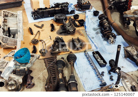 Disassembled engine components spread across workshop floor showing crankshaft, driveshaft, chains, housing and tools. Vehicle check-up and repair. Underbody chassis and suspension parts Disassembled engine components spread across workshop floor showing crankshaft, driveshaft, chains, housing and tools. Vehicle check-up and repair. Underbody chassis and suspension parts 125140007