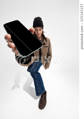 Young man in brown jacket and beanie extends hand with smartphone mockup against white studio background. 125141157