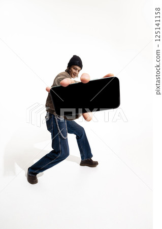 Man in casual streetwear and beanie holds smartphone mockup against white studio background. Man in casual streetwear and beanie holds smartphone mockup against white studio background. 125141158
