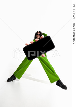 Woman in green with hand extended holding smartphone directly toward viewer against white studio background. 125141161