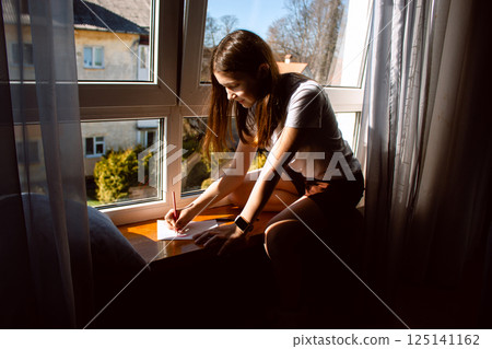 Preteen daughter carefully crafting mother's day, 8 march or birthday card for mom sitting by window at home. Concept of family love, tradition and celebration. Preteen daughter carefully crafting mother's day, 8 march or birthday card for mom sitting by window at home. Concept of family love, tradition and celebration. 125141162