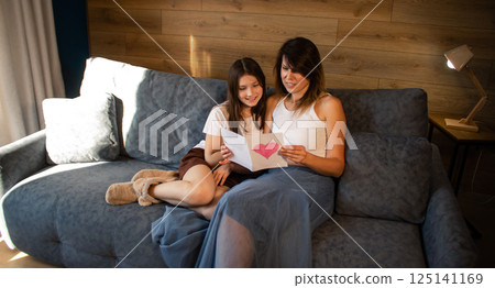 Mom and daughter celebrating mother's day handmade reading greeting card sitting on sofa in home interior. 125141169