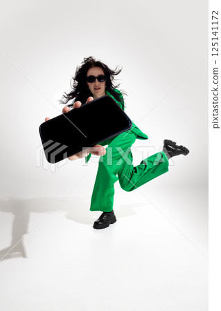 Confident woman leans forward with smartphone with blank screen in one hand, against white studio background. 125141172