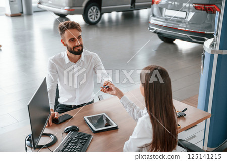 Sitting by the table and taking the keys. Man with woman in white clothes are in the car dealership together Sitting by the table and taking the keys. Man with woman in white clothes are in the car dealership together 125141215