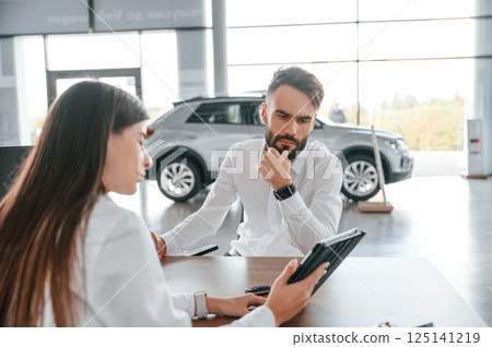 Showing information in the tablet. Sitting by the table. Man with woman in white clothes are in the car dealership Showing information in the tablet. Sitting by the table. Man with woman in white clothes are in the car dealership 125141219