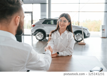 Cheerful customer is smiling. Man with woman in white clothes are in the car dealership together Cheerful customer is smiling. Man with woman in white clothes are in the car dealership together 125141231