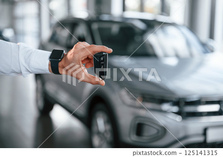 Close up view of keys that are in hand. Young man in white clothes is in the car dealership 125141315