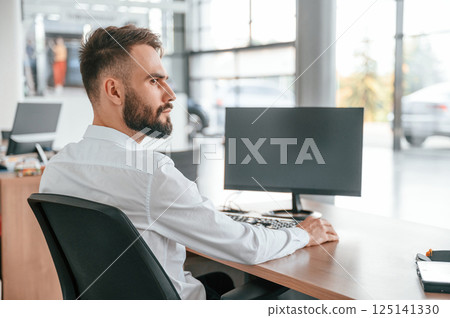 Sitting by the table. Young man in white clothes is in the car dealership 125141330