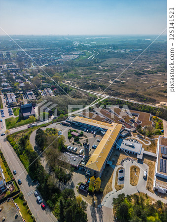 Aerial view of a modern building complex with solar panels, surrounded by residential houses and green areas, located near sandy dunes and walking trails under a clear blue sky. Aerial view of a modern building complex with solar panels, surrounded by residential houses and green areas, located near sandy dunes and walking trails under a clear blue sky. 125141521