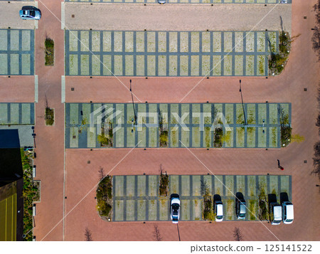 Aerial view of a nearly empty parking lot with a few parked cars, neat rows of green-grid spaces, brick walkways, and young trees casting long shadows in the sunlight. 125141522