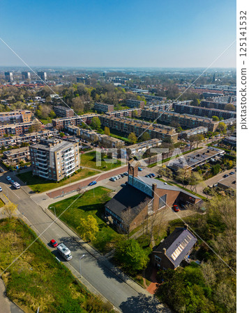 Aerial view of a residential neighborhood with a church at the center, surrounded by apartment buildings, green spaces, roads, and trees under a clear sky. 125141532