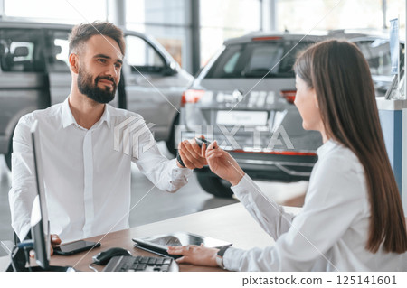 Owner of new automobile is receiving keys. Man with woman in white clothes are in the car dealership together 125141601