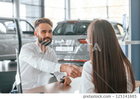 Successful deal. Sitting and doing handshake. Man with woman in white clothes are in the car dealership together Successful deal. Sitting and doing handshake. Man with woman in white clothes are in the car dealership together 125141605