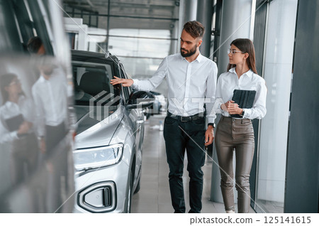 Touching the automobile surface. Man with woman in white clothes are in the car dealership together 125141615