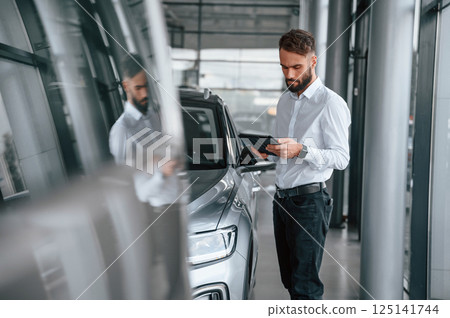 Black graphic tablet in hands. Young man in white clothes is in the car dealership 125141744
