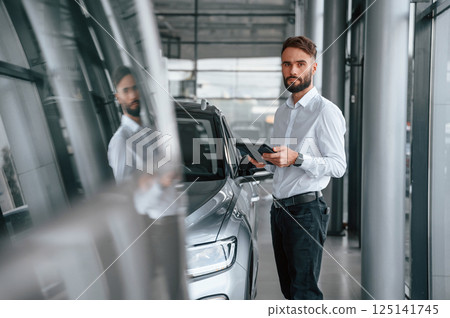 Black graphic tablet in hands. Young man in white clothes is in the car dealership 125141745