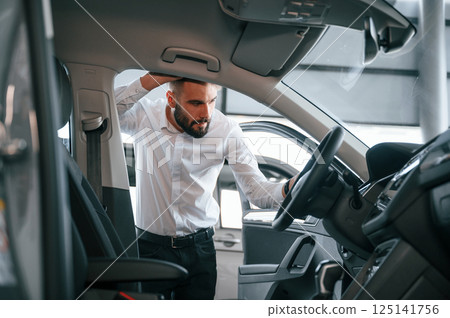 Examining the salon of the automobile. Young man in white clothes is in the car dealership 125141756