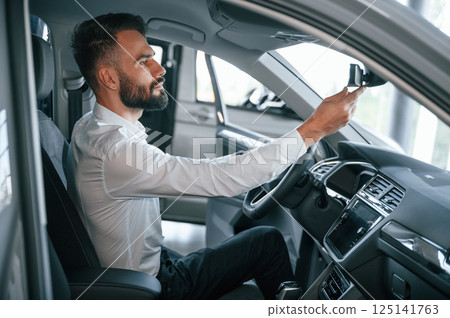 Side view. Sitting inside of a automobile. Young man in white clothes is in the car dealership Side view. Sitting inside of a automobile. Young man in white clothes is in the car dealership 125141763