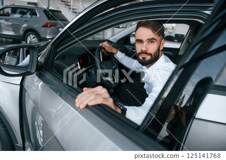 Sitting, looking through the window of the automobile. Young man in white clothes is in the car dealership 125141768