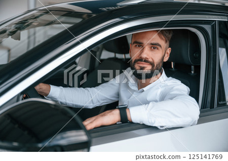 Sitting, looking through the window of the automobile. Young man in white clothes is in the car dealership 125141769