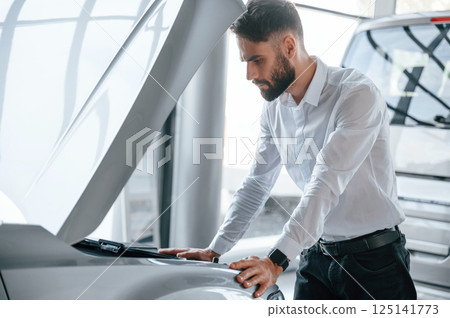 Looks under the hood of automobile. Young man in white clothes is in the car dealership Looks under the hood of automobile. Young man in white clothes is in the car dealership 125141773