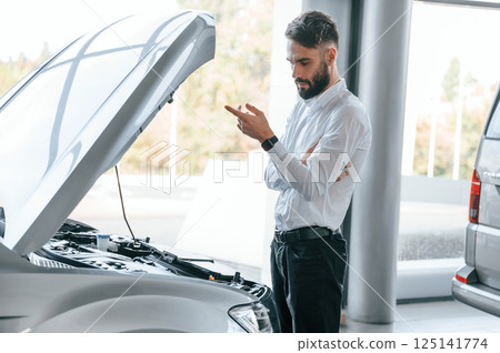 Looks under the hood of automobile. Young man in white clothes is in the car dealership 125141774