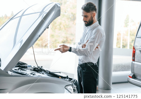 Looks under the hood of automobile. Young man in white clothes is in the car dealership 125141775