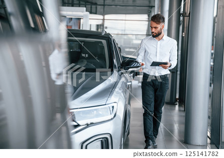Standing and holding black graphic tablet in hand. Young man in white clothes is in the car dealership 125141782