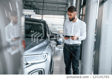 Standing and holding black graphic tablet in hand. Young man in white clothes is in the car dealership 125141783