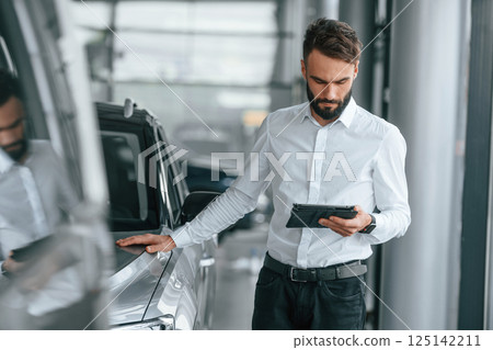 Standing with table in hands. Young man in white clothes is in the car dealership 125142211
