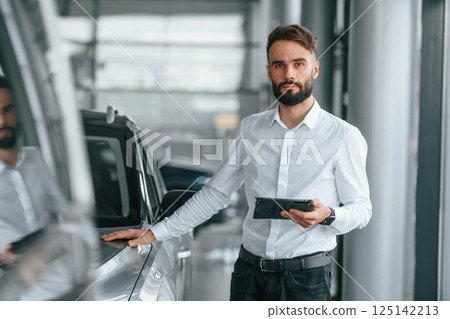 Touching the surface of automobile. Young man in white clothes is in the car dealership Touching the surface of automobile. Young man in white clothes is in the car dealership 125142213