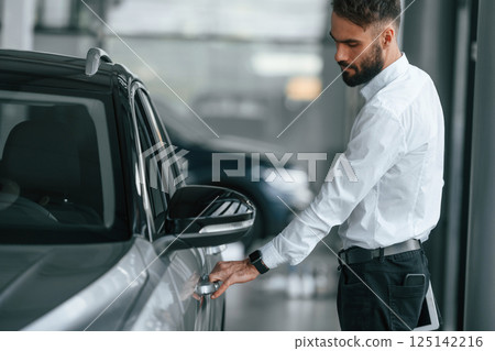 Opening the door. Young man in white clothes is in the car dealership 125142216