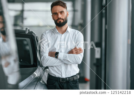 Standing with arms crossed. Young man in white clothes is in the car dealership Standing with arms crossed. Young man in white clothes is in the car dealership 125142218