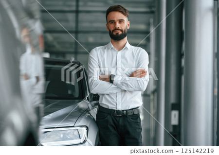 Standing with arms crossed. Young man in white clothes is in the car dealership 125142219