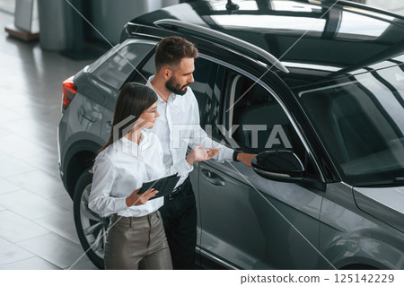 Assistant is helping the customer. Man with woman in white clothes are in the car dealership together 125142229