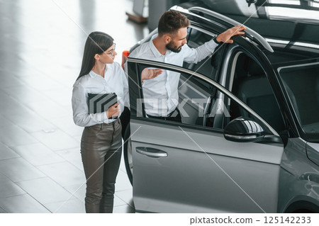 Opening the door. Man with woman in white clothes are in the car dealership together 125142233