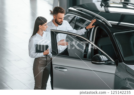 Opening the door. Man with woman in white clothes are in the car dealership together 125142235