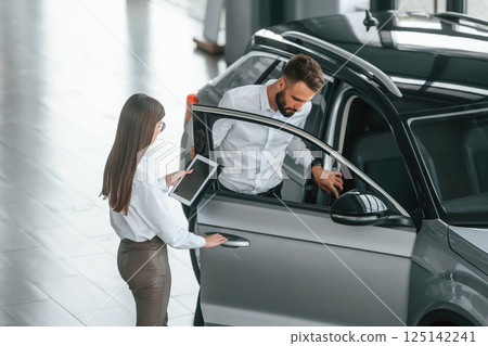 Opening the door. Man with woman in white clothes are in the car dealership together 125142241