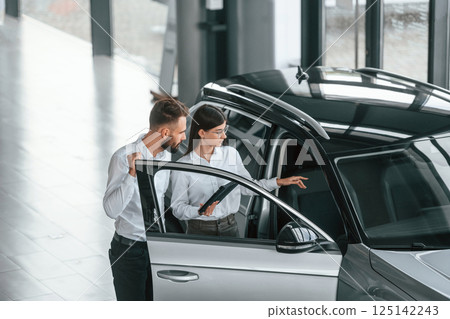 Door is opened. Man with woman in white clothes are in the car dealership together 125142243