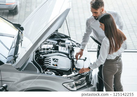 What's under the hood. Man with woman in white clothes are in the car dealership together What's under the hood. Man with woman in white clothes are in the car dealership together 125142255