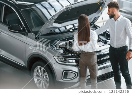 What's under the hood. Man with woman in white clothes are in the car dealership together What's under the hood. Man with woman in white clothes are in the car dealership together 125142256