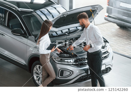 Hood is opened. Man with woman in white clothes are in the car dealership together 125142260