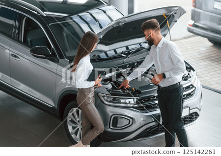 Hood is opened. Man with woman in white clothes are in the car dealership together Hood is opened. Man with woman in white clothes are in the car dealership together 125142261