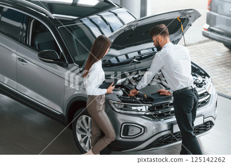 Hood is opened. Man with woman in white clothes are in the car dealership together 125142262
