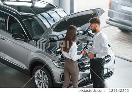Hood is opened. Man with woman in white clothes are in the car dealership together Hood is opened. Man with woman in white clothes are in the car dealership together 125142263