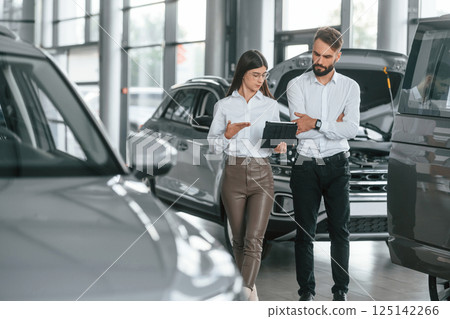 Reading information by using tablet. Man with woman in white clothes are in the car dealership together 125142266