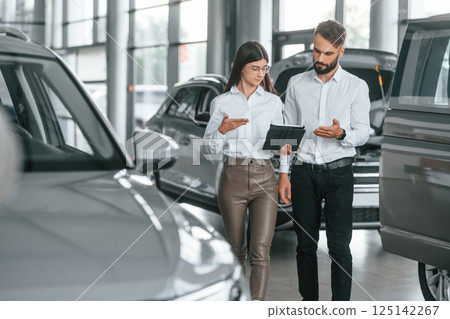 Reading information by using tablet. Man with woman in white clothes are in the car dealership together 125142267